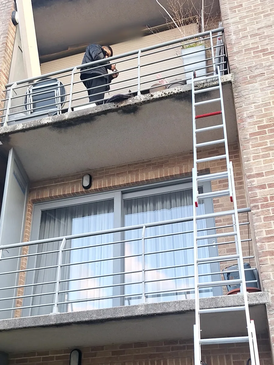 Échelle posée depuis le jardin jusqu'au balcon du deuxième étage, où le technicien intervient sur le volet roulant descendu.
