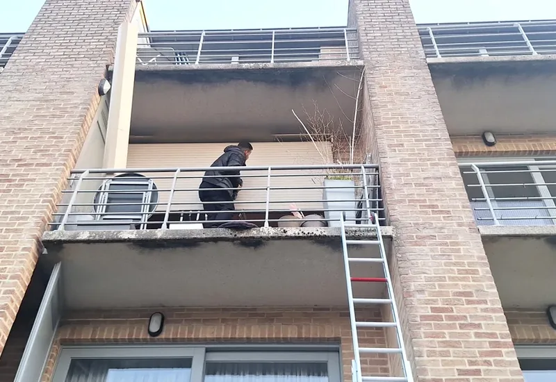 Technicien sur le balcon du deuxième étage, devant un volet roulant électrique bloqué en position basse, avec une échelle depuis le jardin, à Auderghem