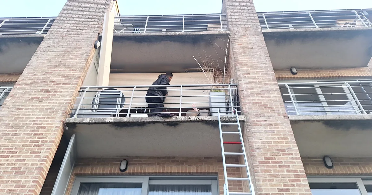 Technicien sur le balcon du deuxième étage, devant un volet roulant électrique bloqué en position basse ; une échelle monte depuis le jardin. Immeuble à Auderghem, secteur Hankar.